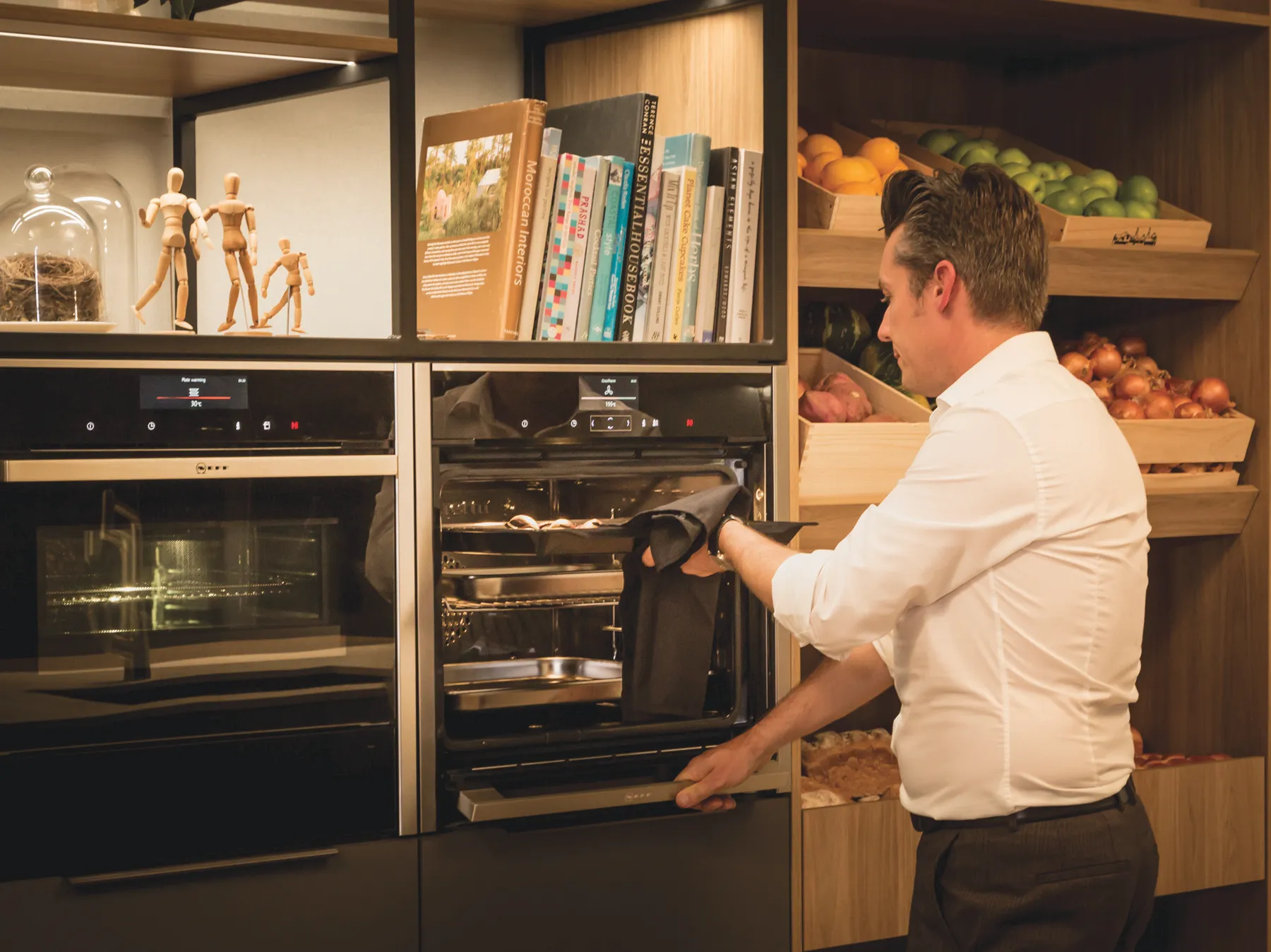 A man in a white shirt carefully removes a tray of cookies or small pastries from an open NEFF built-in oven, using a black cloth. To his left, another NEFF oven displays '30°C' on its digital screen. 