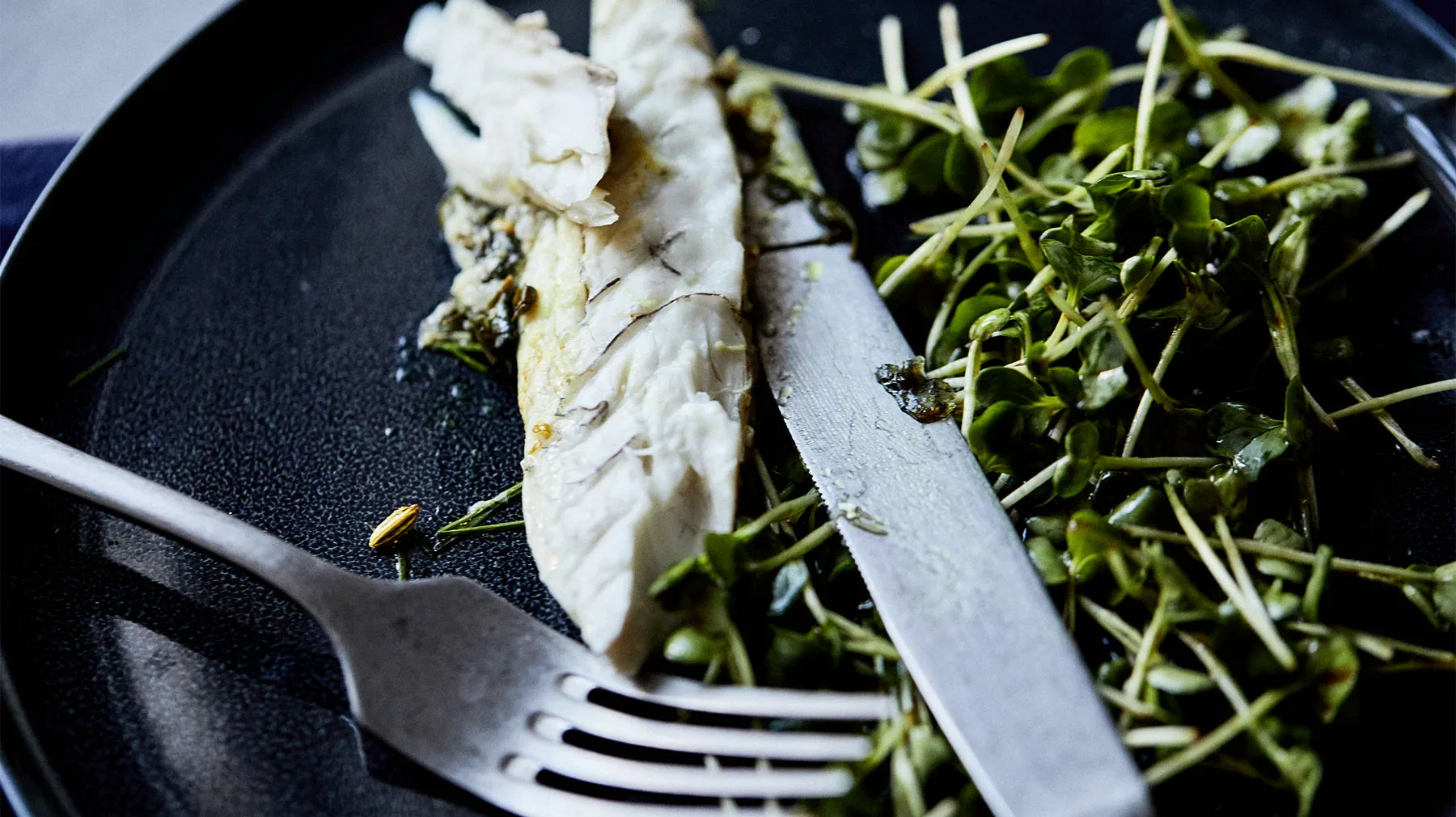 Piatto con pesce alla griglia, verdi freschi e utensili su uno sfondo scuro.