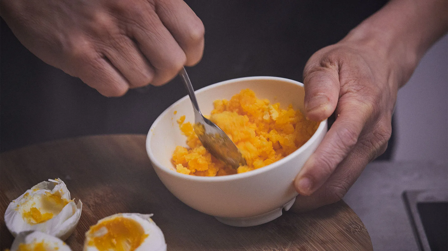 Hands mashing cooked yolks in a bowl on a wooden table, with peeled eggs nearby.
