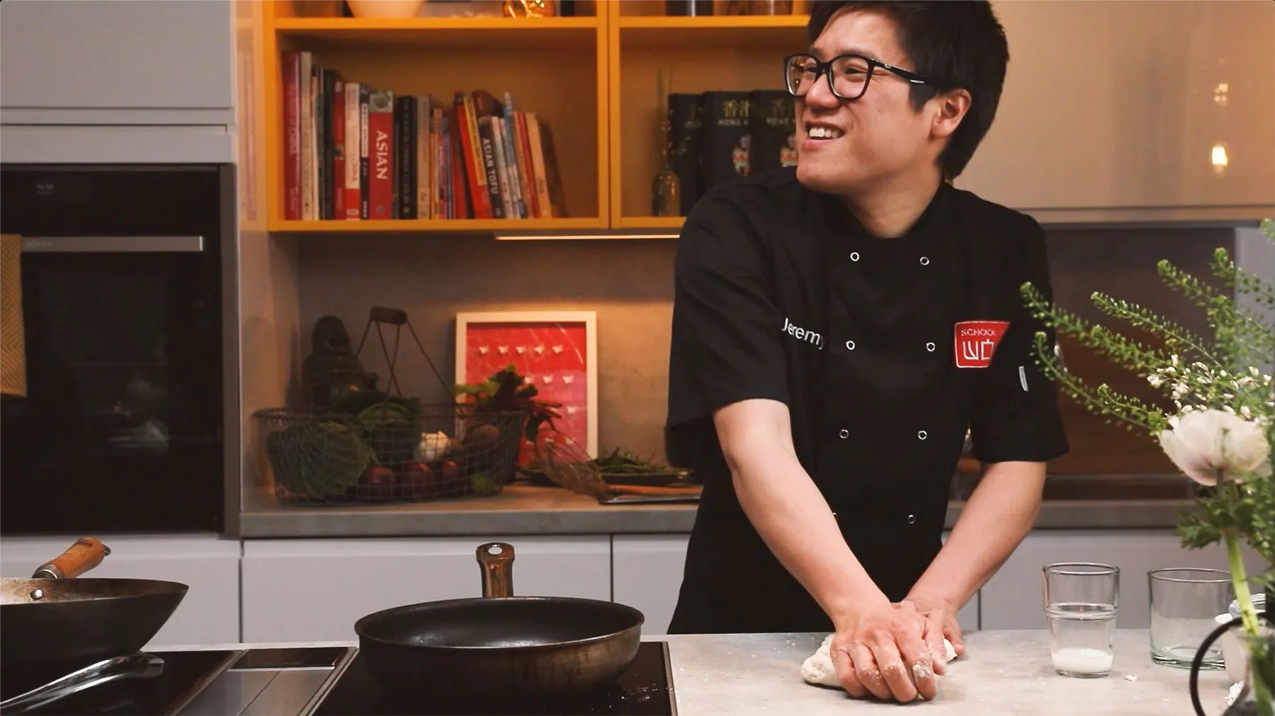 A smiling young man in a chef's uniform stands in a kitchen with bookshelves in the background.