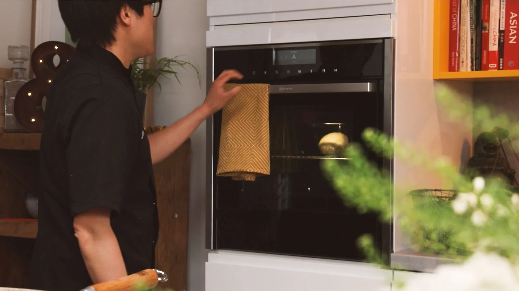Person opening oven door in kitchen, with books and plants visible in background.