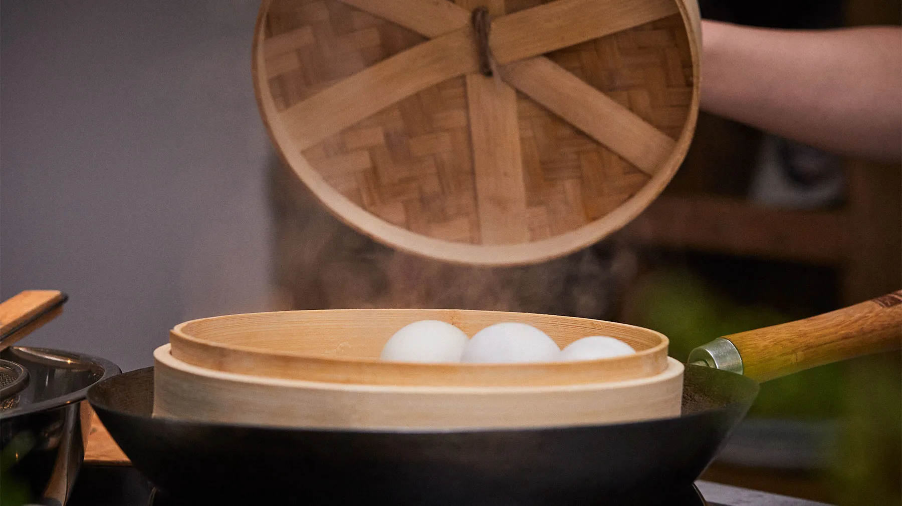 Wooden steamer with dough, cooking utensils, and a round wooden lid in a kitchen setting.