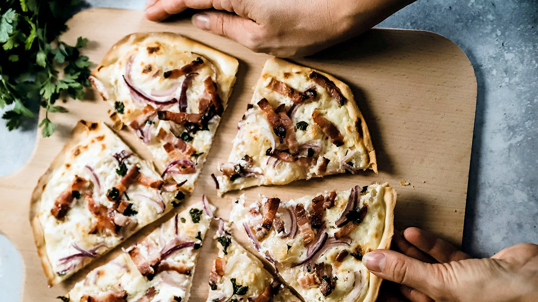 Flammkuchen mit Speck und Zwiebeln auf einem Holzbrett, zwei Hände greifen danach