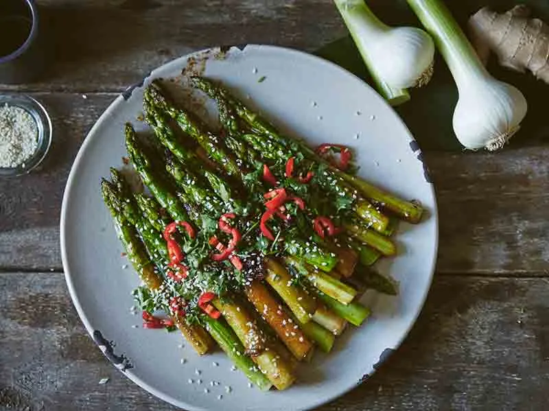 O farfurie cu sparanghel la grătar, garnisit cu ardei iute roșu, ierburi aromatice și semințe de susan, alături de ceapă verde proaspătă și ghimbir pe o masă de lemn.