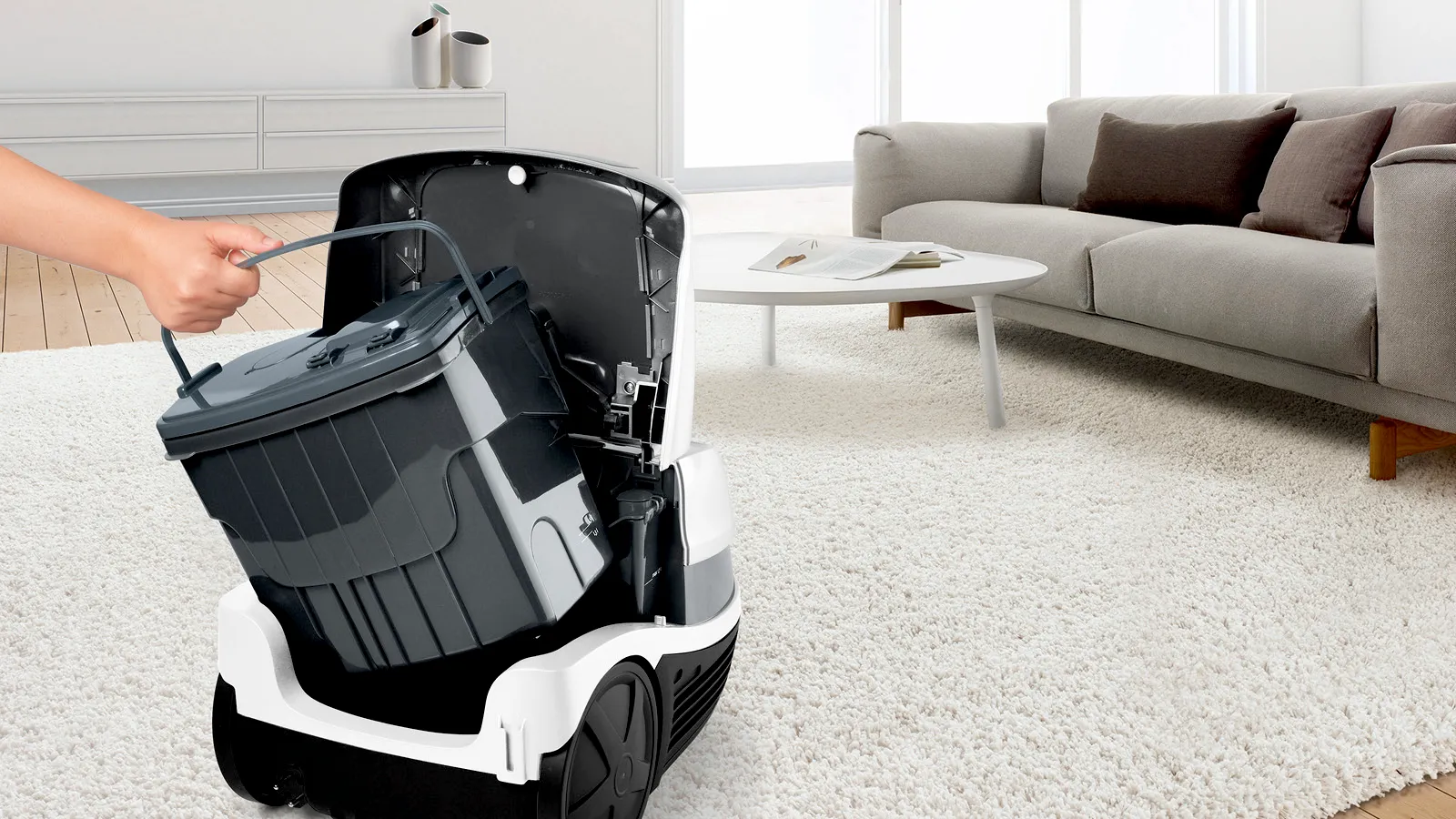 O mână scoate un coș de gunoi negru dintr-un aspirator robot alb, cu capacul deschis, într-un living modern.