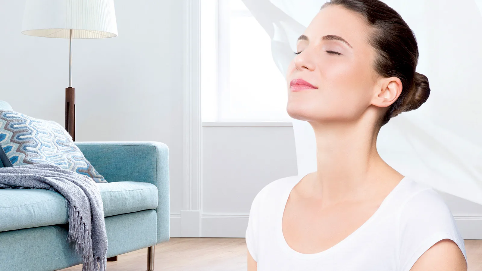 O femeie cu ochii închiși, care se bucură de aerul proaspăt sau de momente de relaxare într-un living luminos și modern, cu o canapea albastru deschis și o lampă de podea.