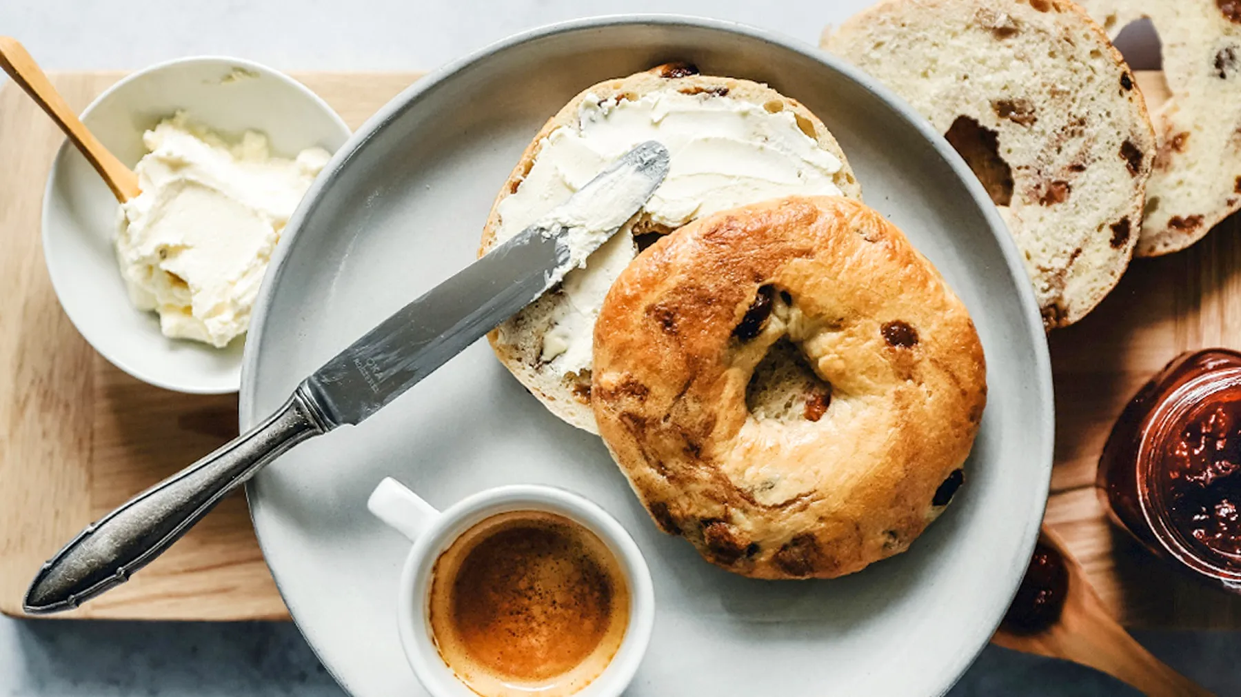 Bagel mit Frischkäse auf grauem Teller neben Espresso, Butter und Marmelade auf Holztisch