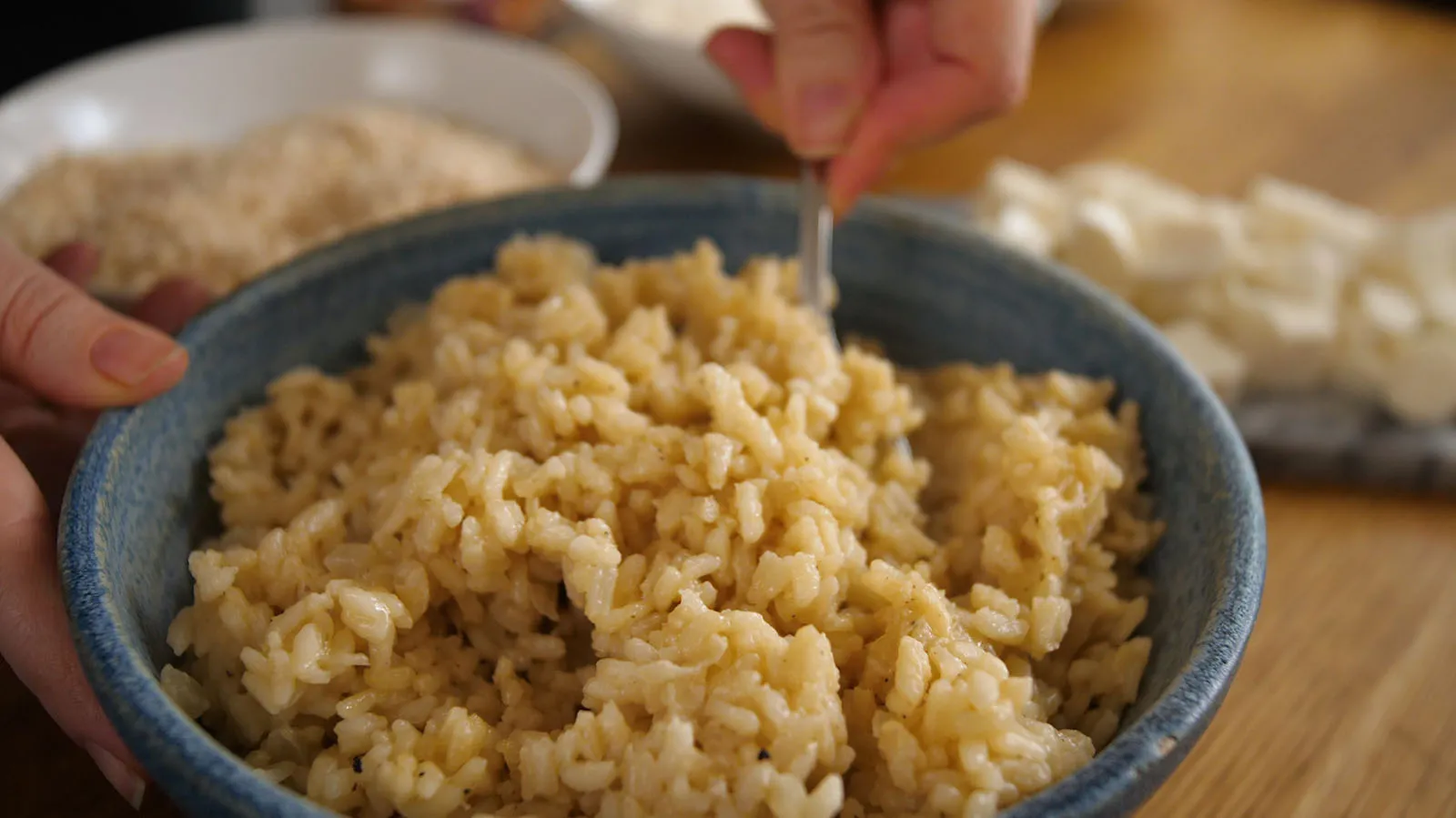 A close-up of a blue bowl filled with cooked brown rice, being stirred with a fork, with diced mozzarella and breadcrumbs visible in the background.