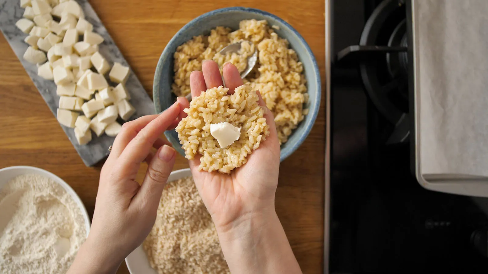 An overhead shot of hands shaping a rice ball with a small piece of mozzarella cheese in the center, surrounded by bowls of cooked rice, flour, breadcrumbs, and diced mozzarella.