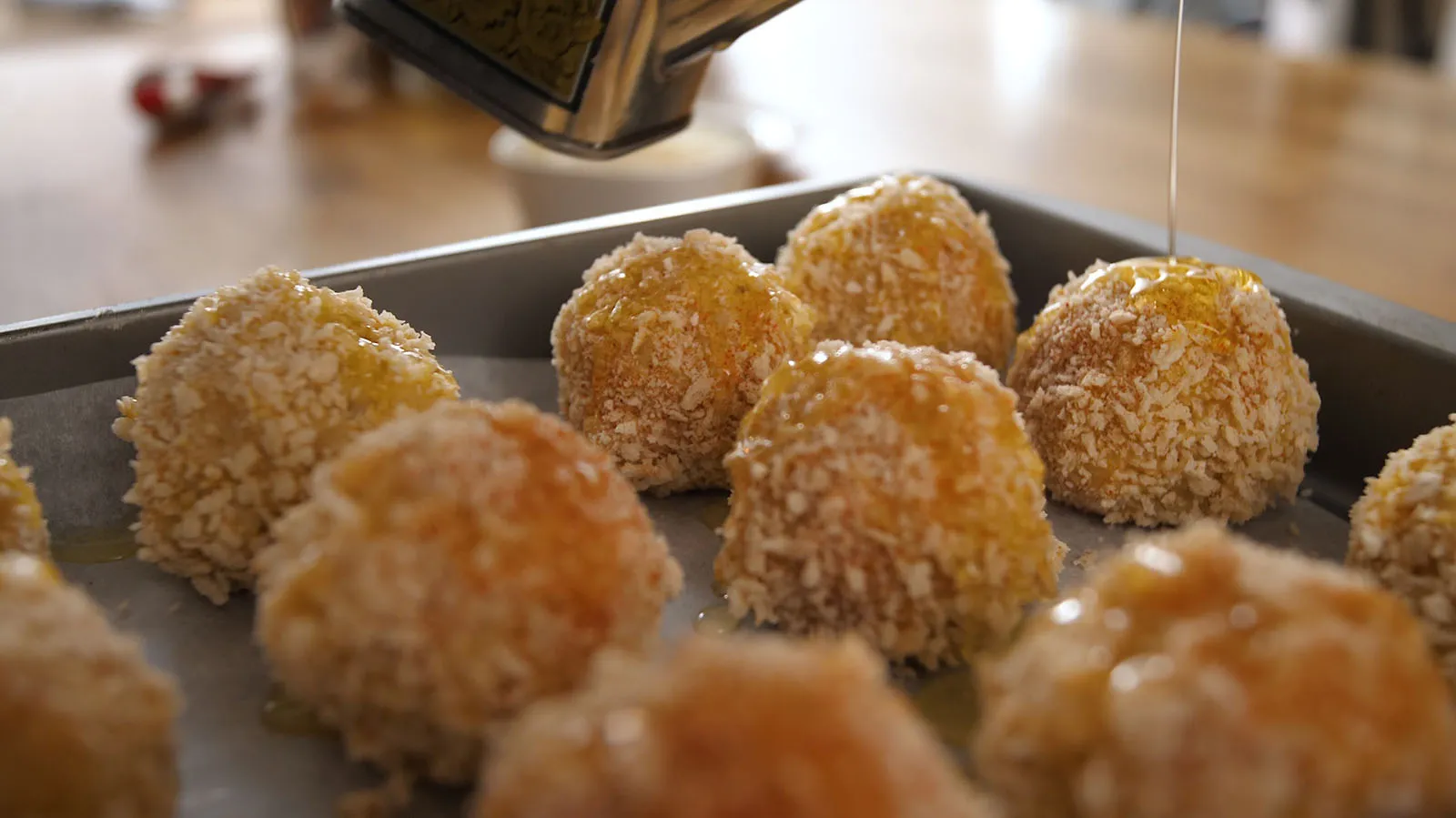 A close-up of golden-brown, breaded rice balls arranged on a baking sheet lined with parchment paper, being drizzled with honey or oil.