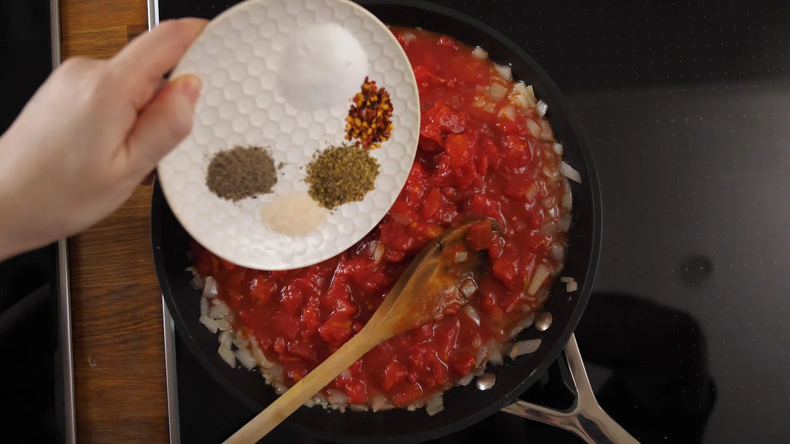 A hand holds a white plate with various spices (salt, pepper, garlic powder, dried herbs, chili flakes) over a black frying pan containing diced tomatoes and onions, ready to be added to the sauce.