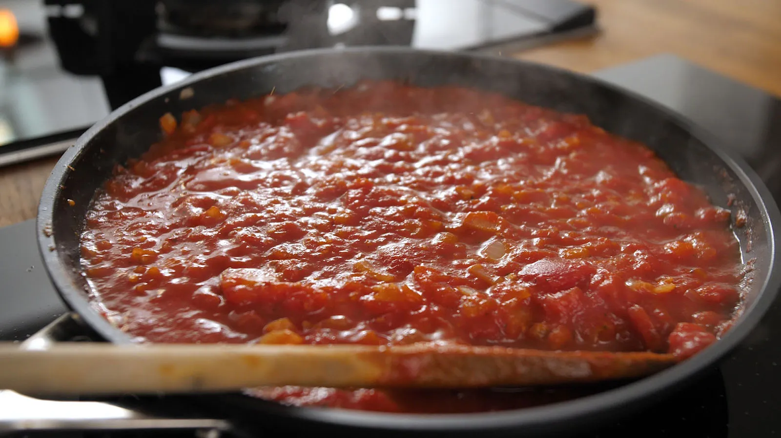 A close-up of a simmering red tomato sauce with diced vegetables in a black frying pan on an induction cooktop, being stirred with a wooden spoon.
