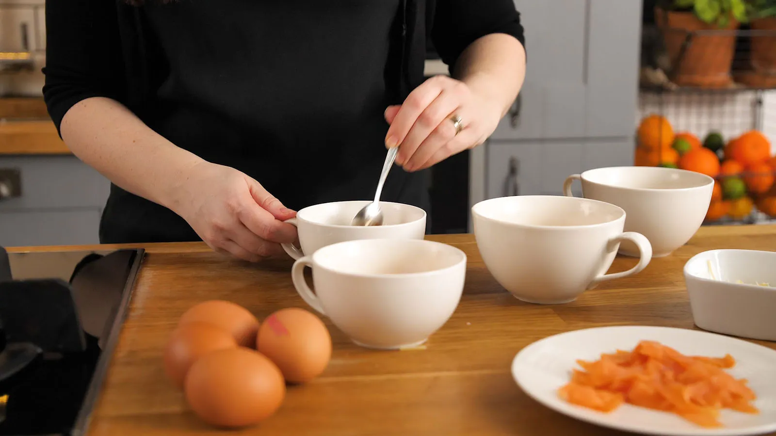A woman prepares several white mugs on a wooden countertop, stirring one with a spoon, with brown eggs and smoked salmon nearby.