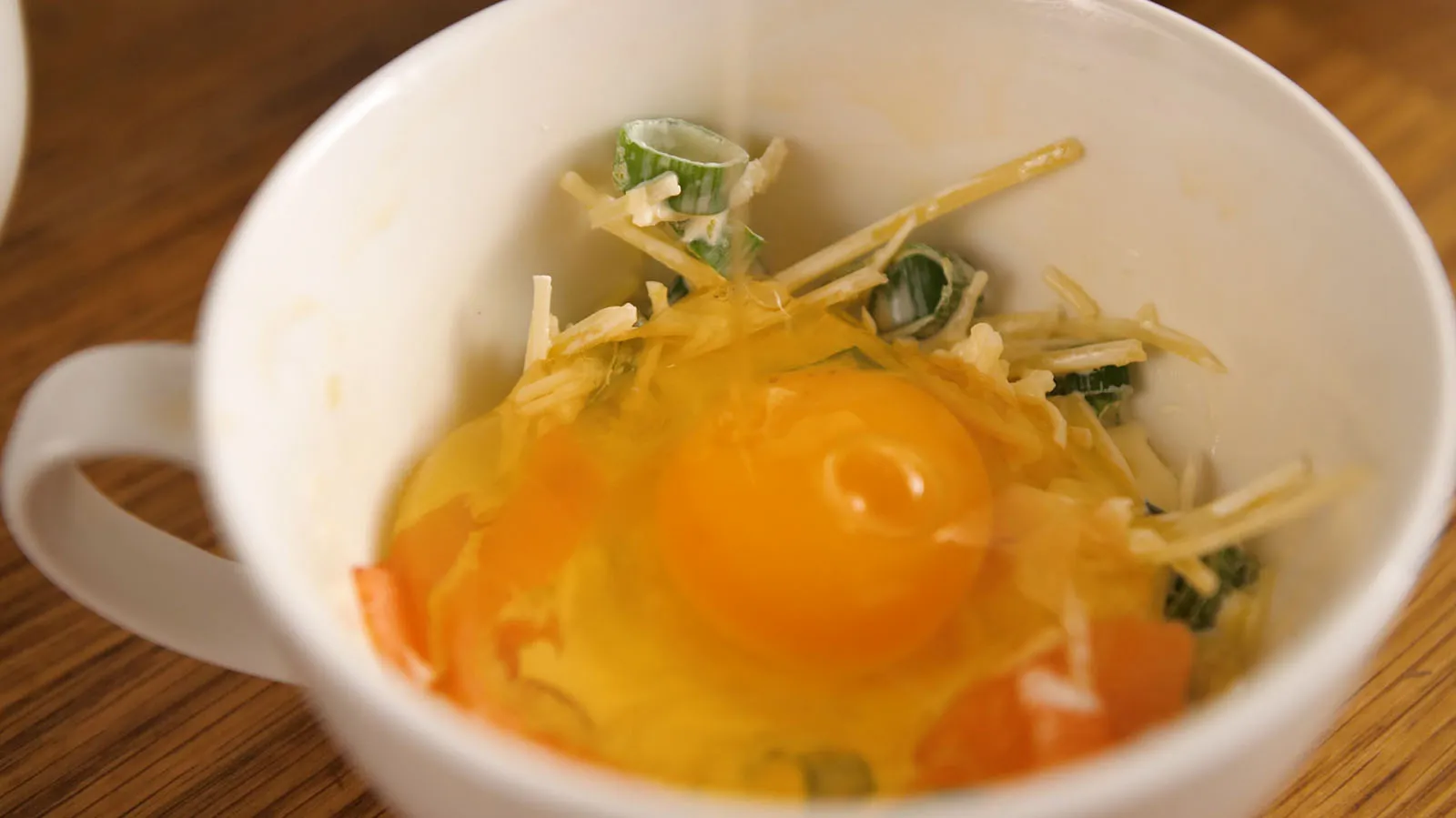 A close-up of a white mug containing a cracked raw egg over shredded cheese and chopped green onions.