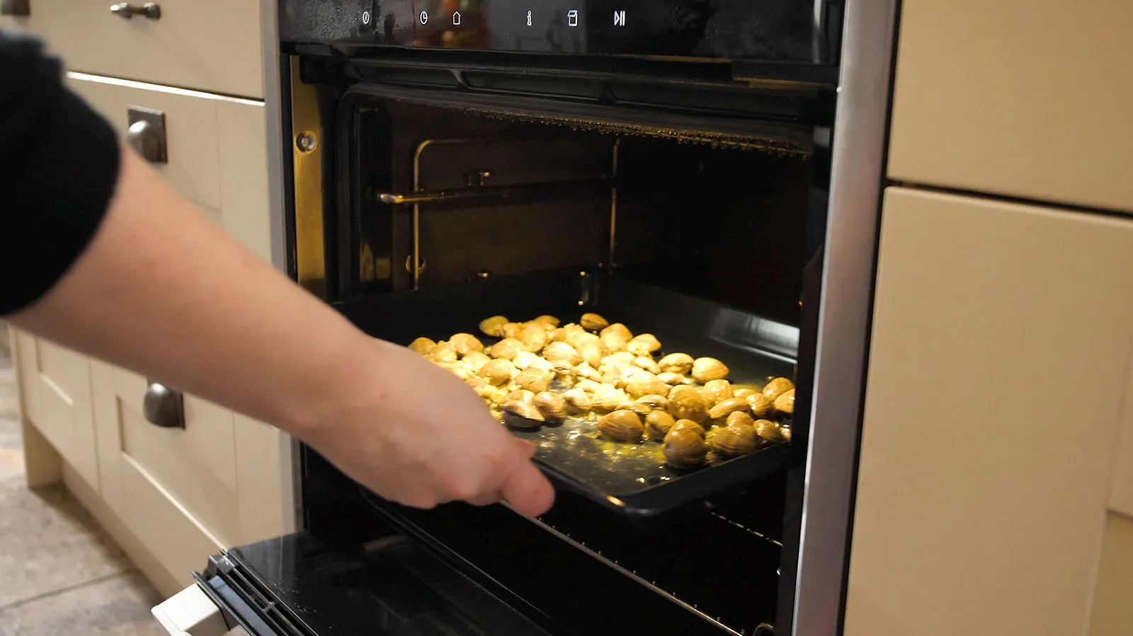 A hand slides a black baking tray filled with cooked clams into a hot oven.