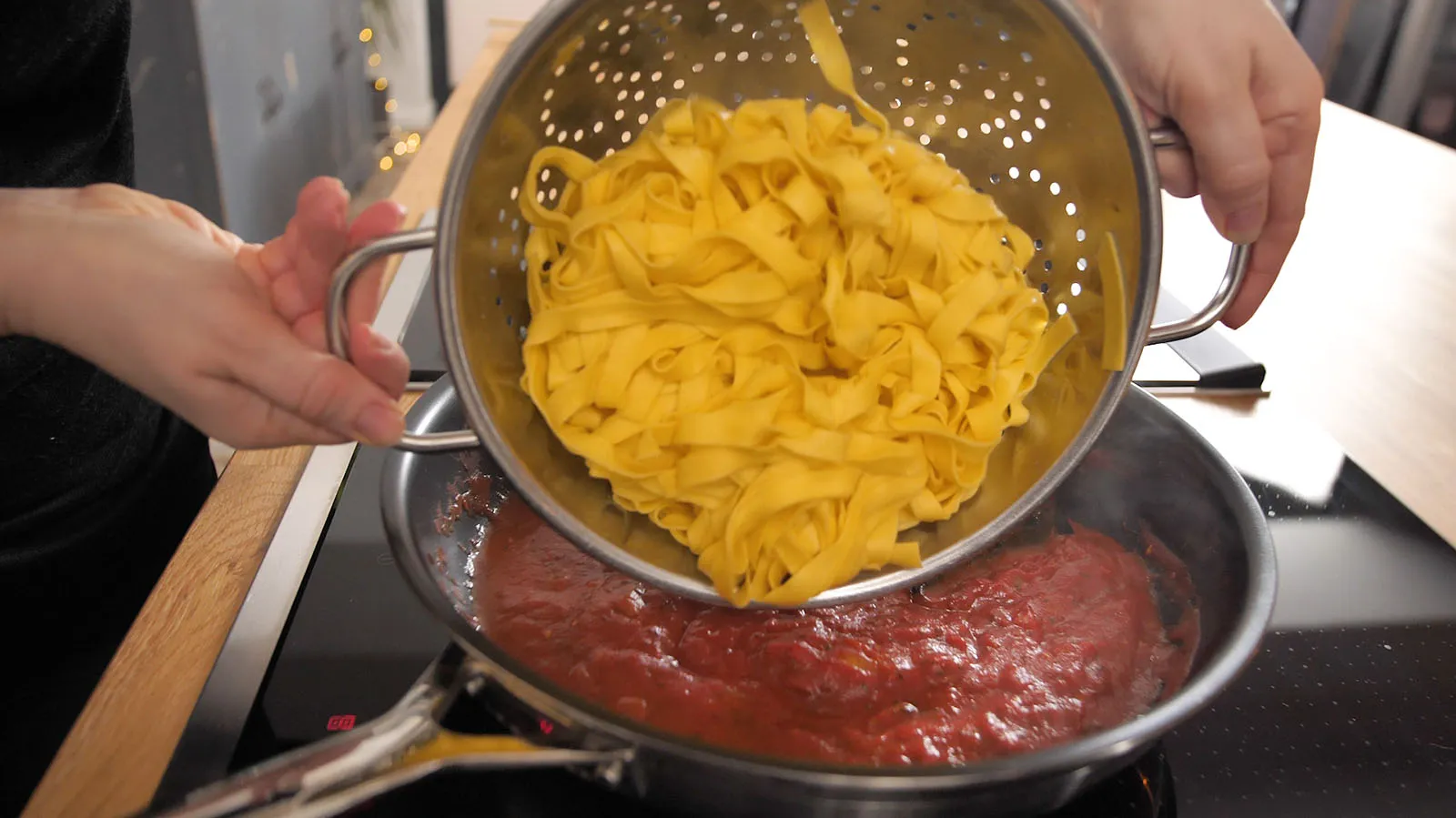 Hands hold a metal colander filled with cooked yellow tagliatelle pasta, draining it over a frying pan of red sauce on an induction cooktop.