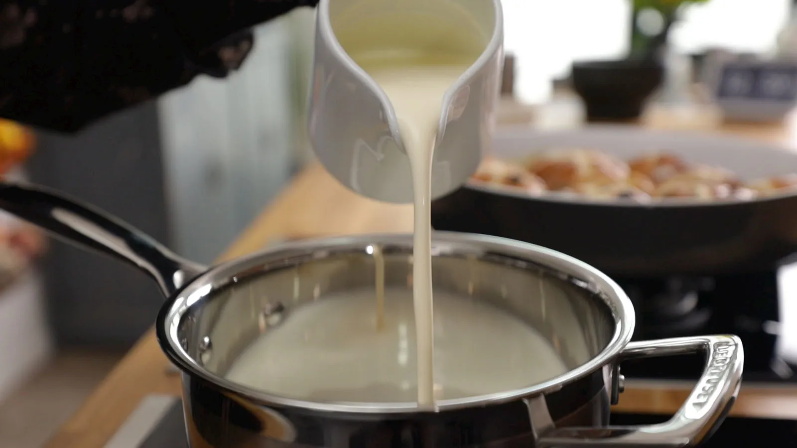 A hand pours white liquid (likely cream or milk) from a white pitcher into a stainless steel saucepan on an induction cooktop.