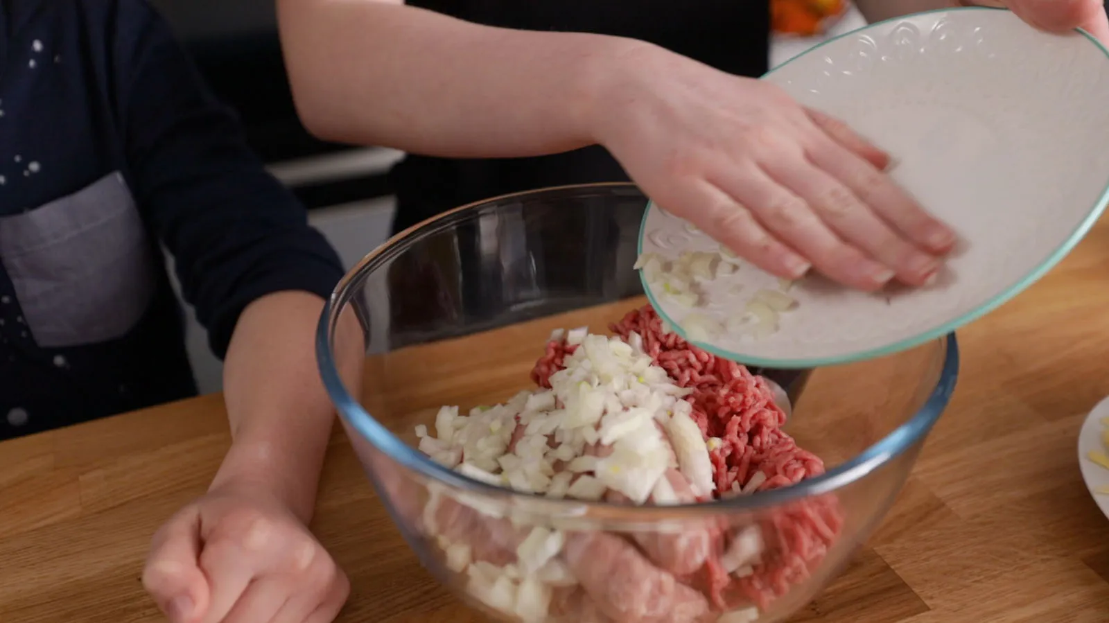 Hands pour diced onions from a white plate into a large glass bowl containing ground meat, preparing meatballs.
