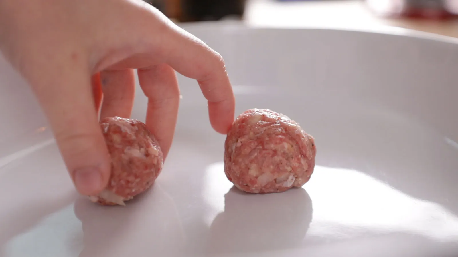 A hand places a raw, round meatball into a white baking dish, next to another raw meatball.