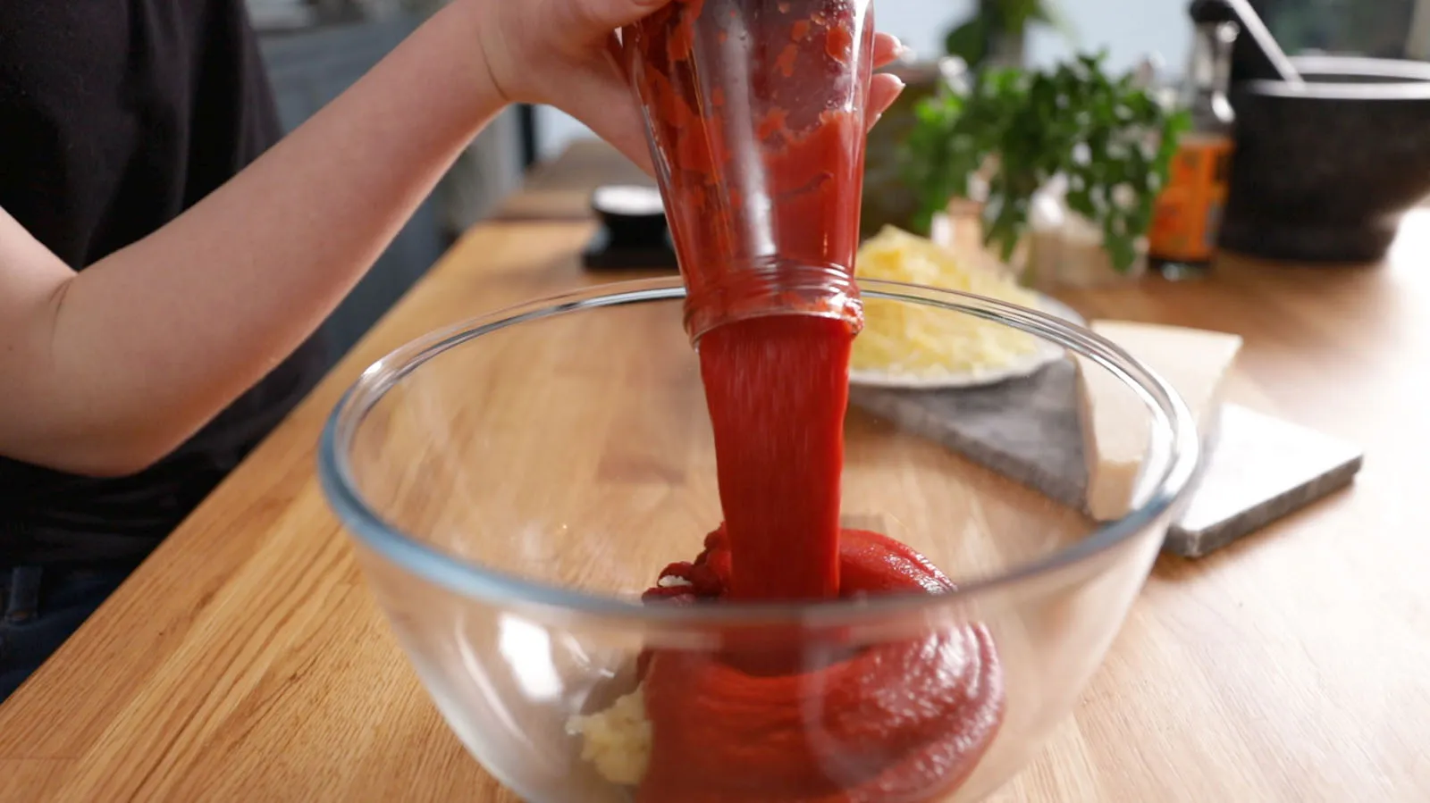 A hand pours thick red tomato paste from a jar into a clear glass bowl containing minced garlic, preparing a sauce.