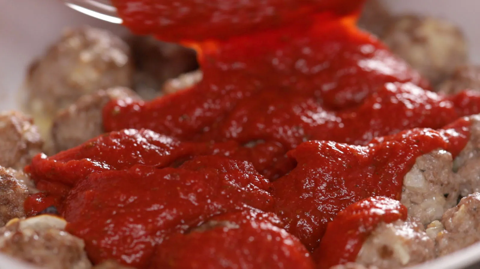 A close-up of a white baking dish where a rich red tomato sauce is being poured over browned meatballs.