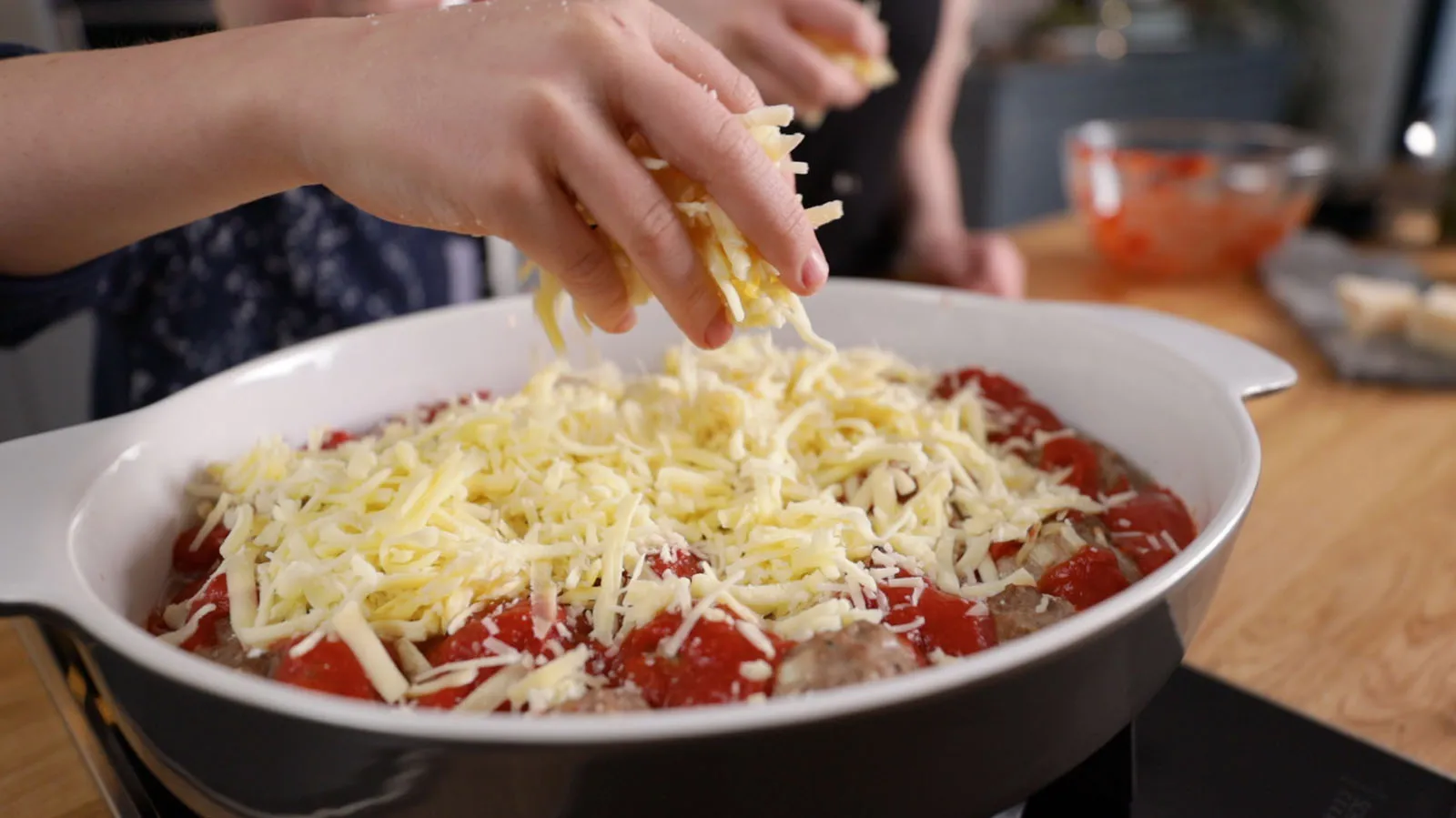 Hands sprinkle shredded cheese over a white baking dish filled with meatballs and diced tomatoes, ready for baking.