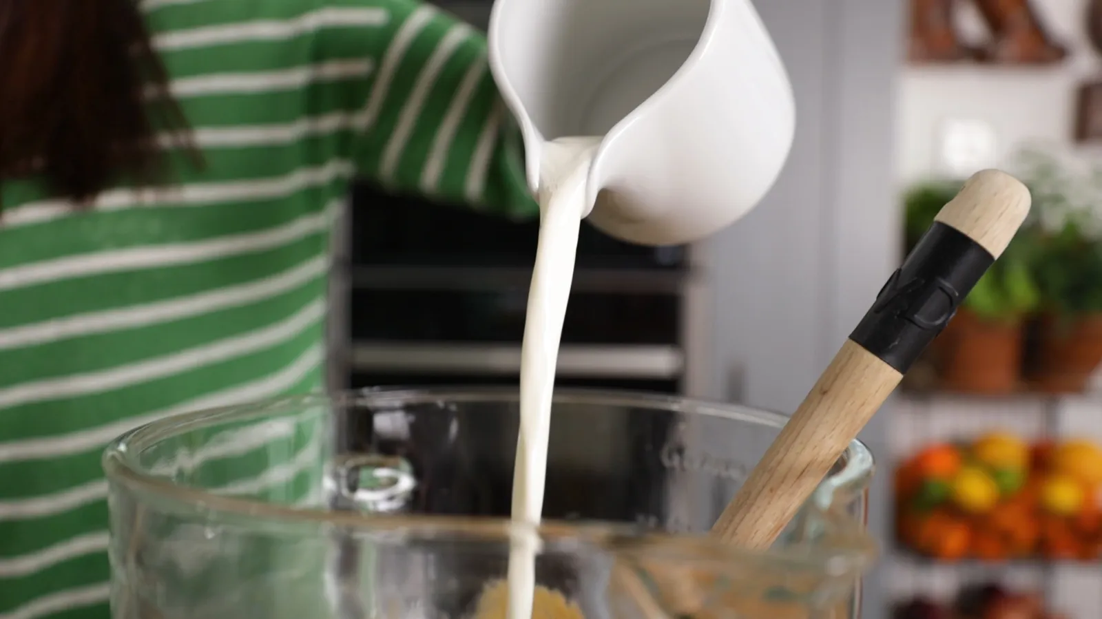 A hand pours white liquid (likely milk or cream) from a white pitcher into a large clear glass mixing bowl.