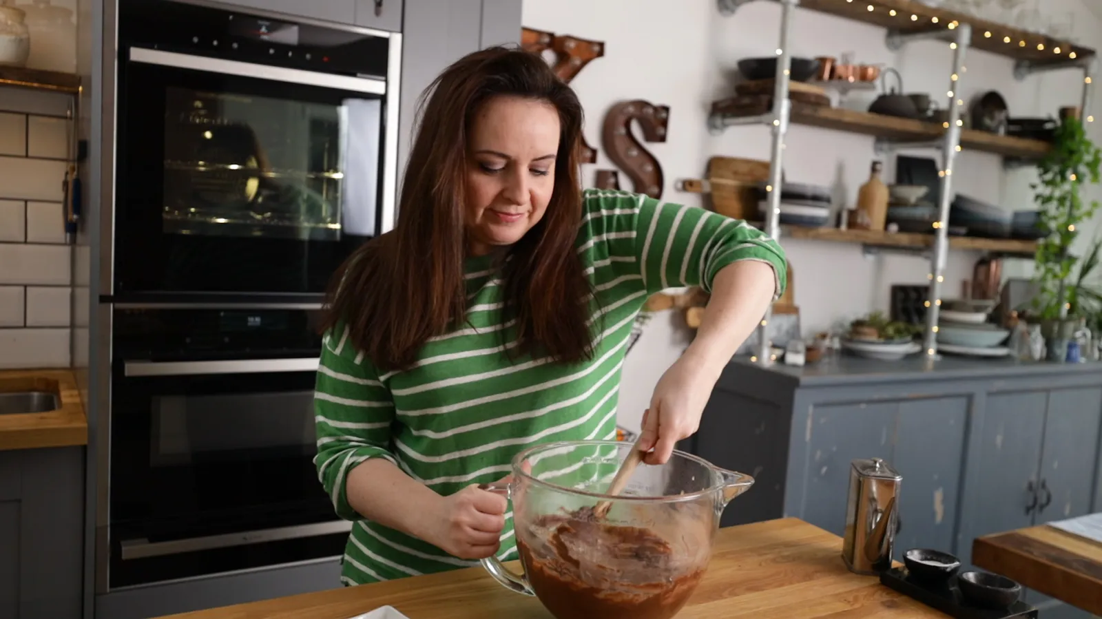 A woman in a green and white striped sweater mixes dark chocolate cake batter in a large clear glass bowl with a wooden spoon in a kitchen.