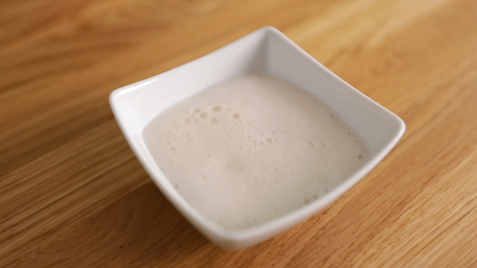 A close-up of a white square bowl filled with foamy white liquid, possibly milk or egg whites.