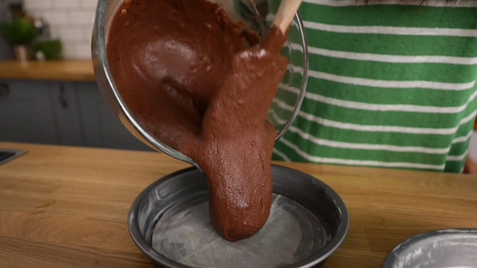A hand pours thick chocolate cake batter from a clear glass bowl into a round cake pan lined with parchment paper.