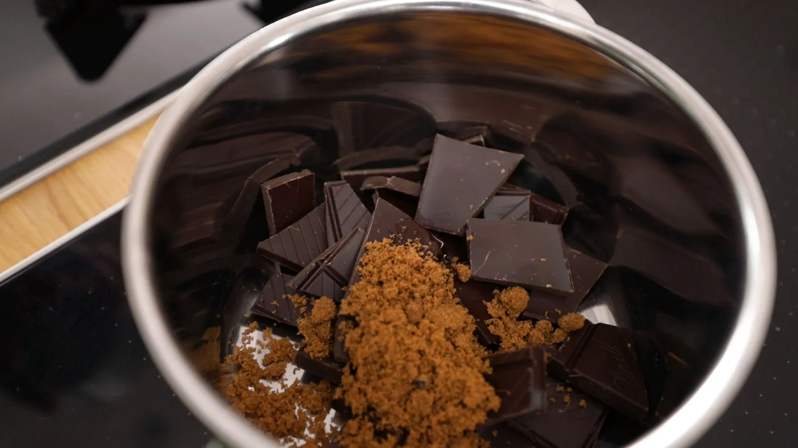 An overhead view of a stainless steel saucepan containing dark chocolate pieces and brown sugar, ready to be melted.