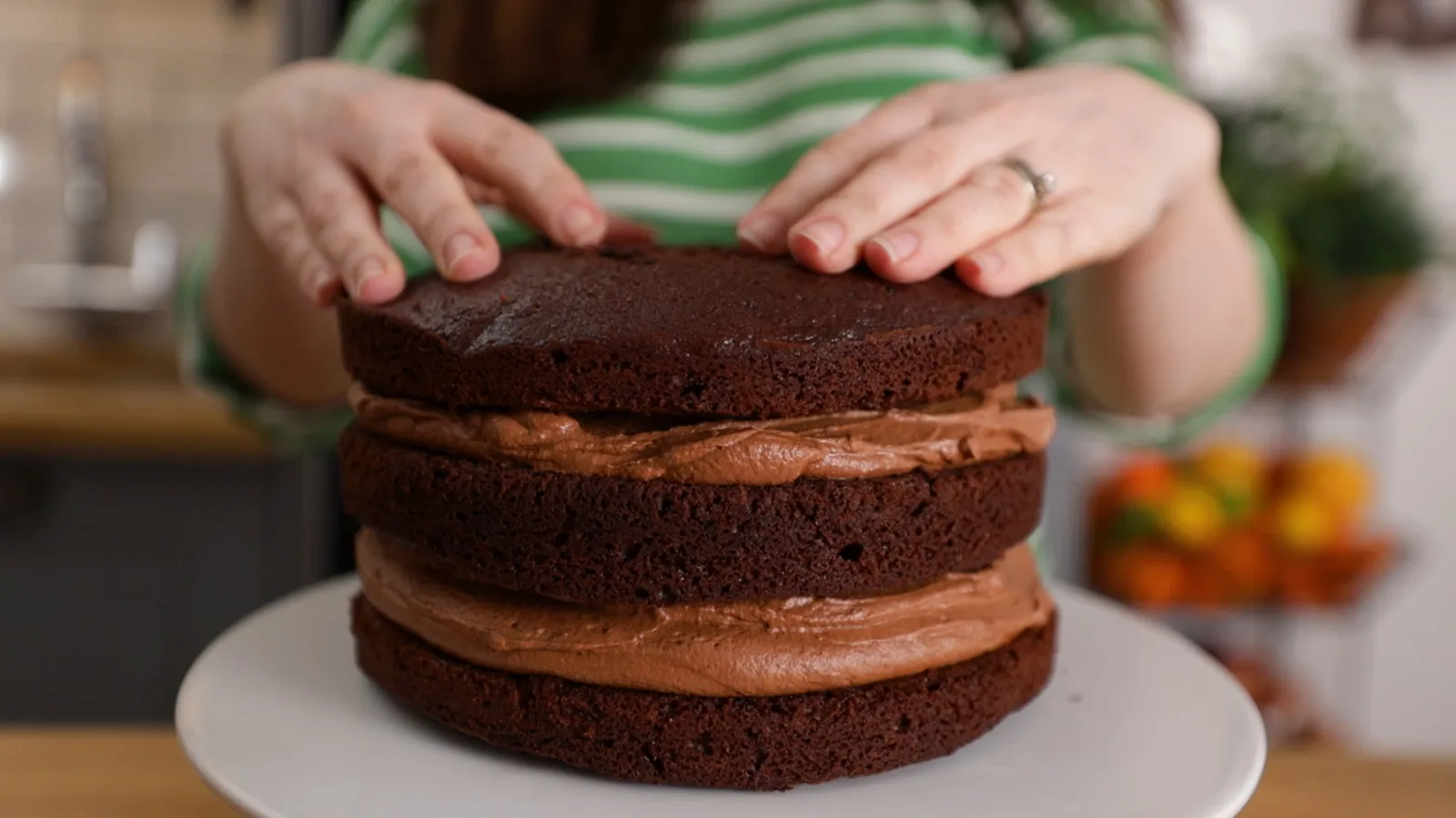 Hands carefully place a chocolate cake layer on top of a layer of chocolate frosting, assembling a multi-layered cake on a white cake stand.