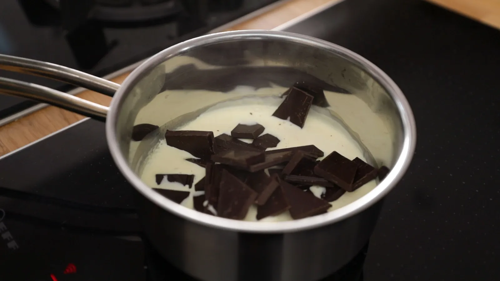 An overhead view of a stainless steel saucepan on an induction cooktop, containing white cream and dark chocolate pieces, melting together.