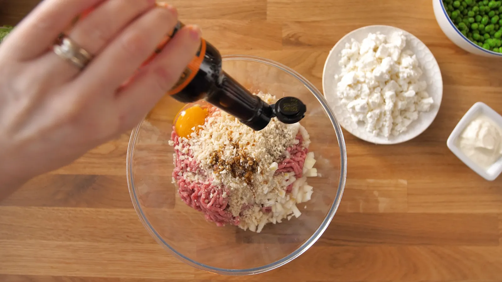 A hand pours a dark liquid (likely soy sauce) from a bottle into a glass bowl containing ground meat, breadcrumbs, and an egg yolk.