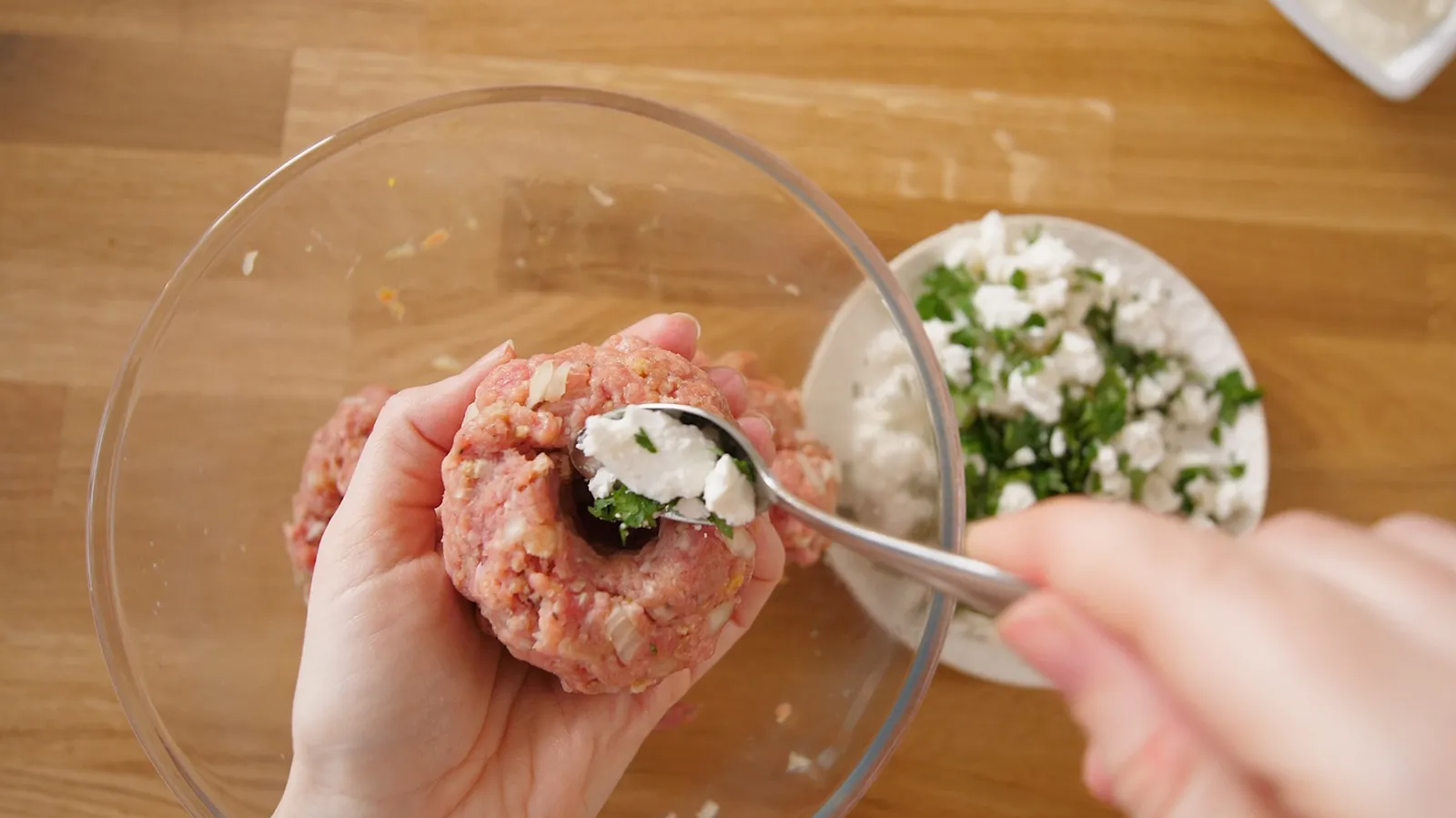 Hands use a spoon to fill the center of a raw burger patty with white crumbled feta cheese and green herbs.