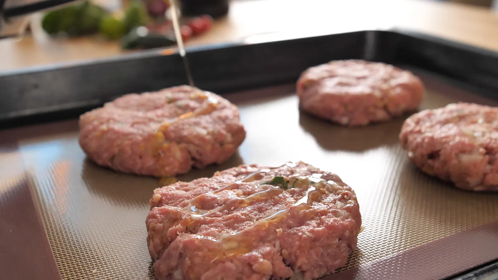 A close-up of four raw burger patties on a baking sheet lined with parchment paper, being drizzled with olive oil.