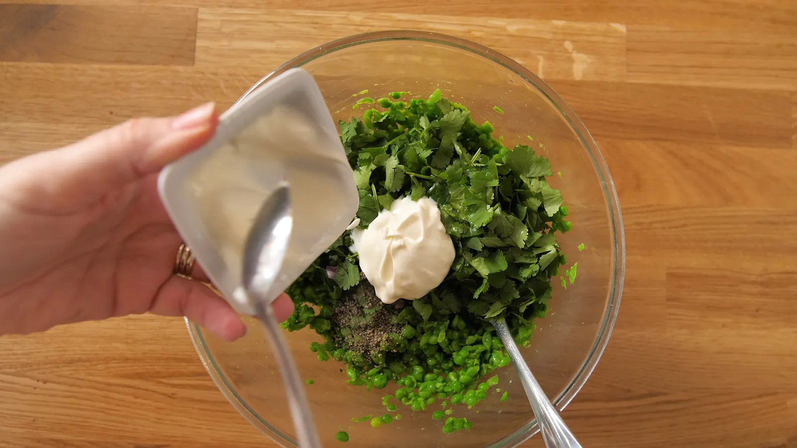 An overhead view of a hand adding a spoonful of white dressing to a glass bowl containing mashed green peas and chopped cilantro.