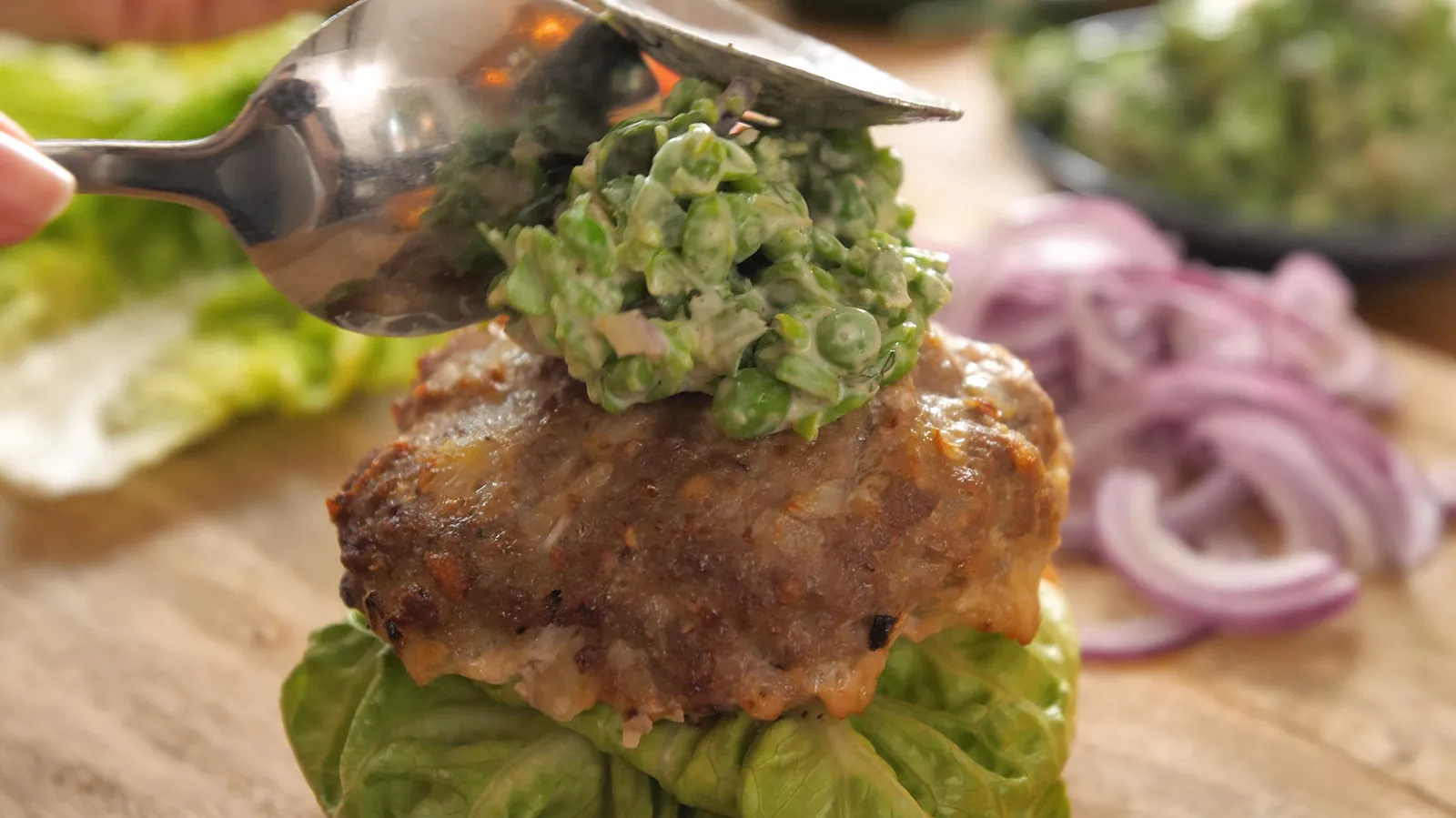 A hand uses a spoon to dollop a green pea and feta mash onto a cooked burger patty, which rests on a bed of lettuce.