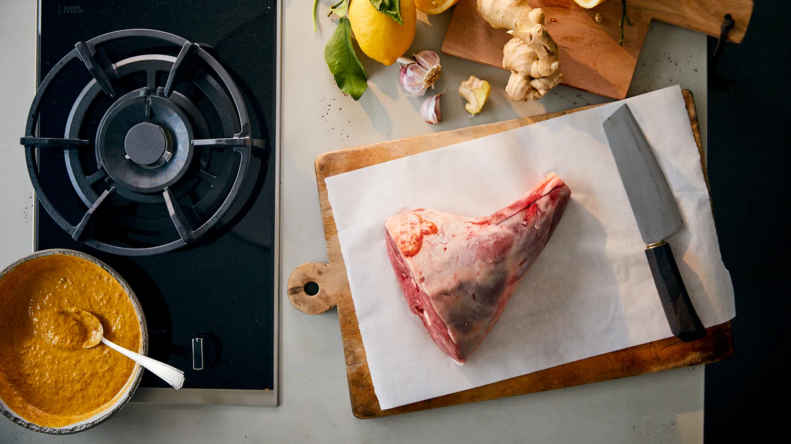 An overhead view of a raw lamb leg on a wooden cutting board with parchment paper, next to a large knife, a gas hob, and a bowl of yellow marinade.