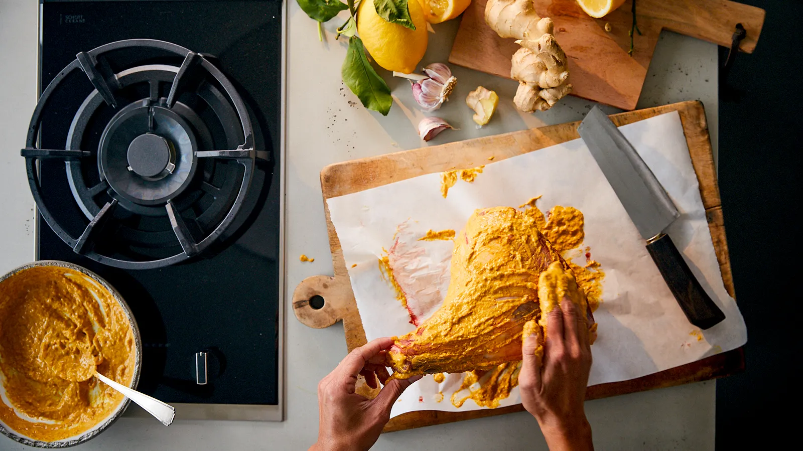 An overhead view of hands coating a raw lamb leg with a yellow marinade on a wooden cutting board, next to a gas hob and various ingredients like lemons, ginger, and garlic.
