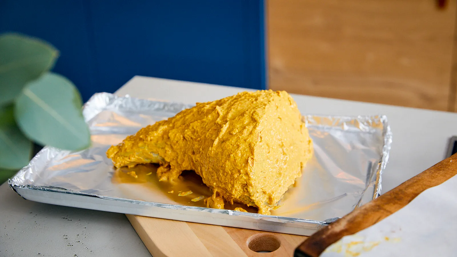 A raw lamb leg coated in a thick yellow marinade, resting on a foil-lined baking tray on a wooden cutting board.