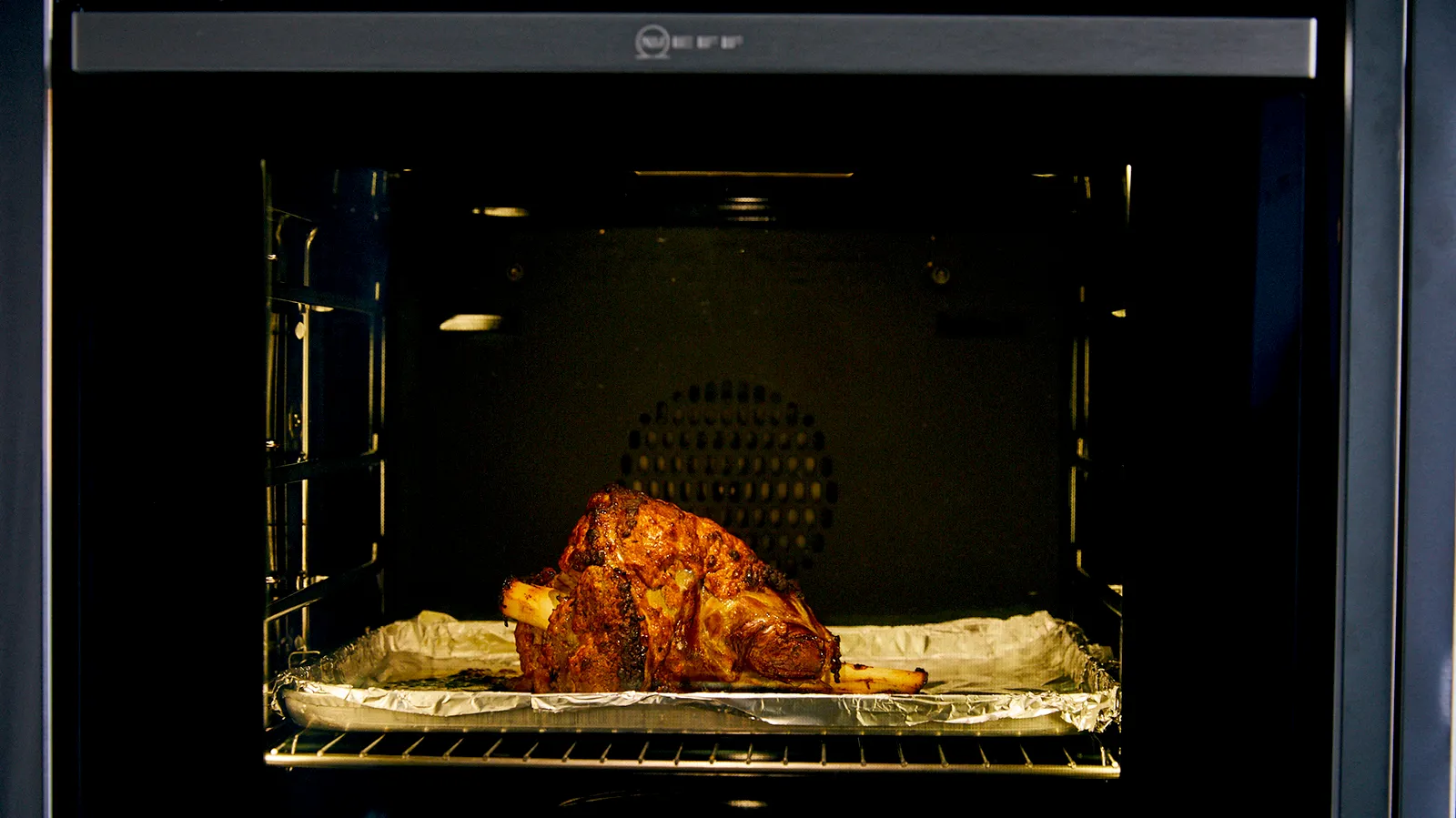 A dark oven interior with a roasted lamb leg on a foil-lined baking tray, showing its crispy brown skin.