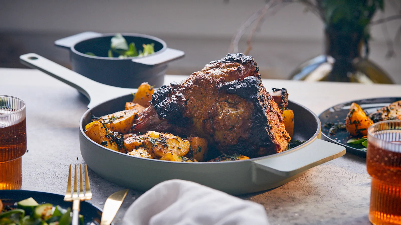 A roasted lamb leg with crispy skin and golden potatoes, garnished with herbs, served in a light green pan, with side salads and drinks on a grey table.