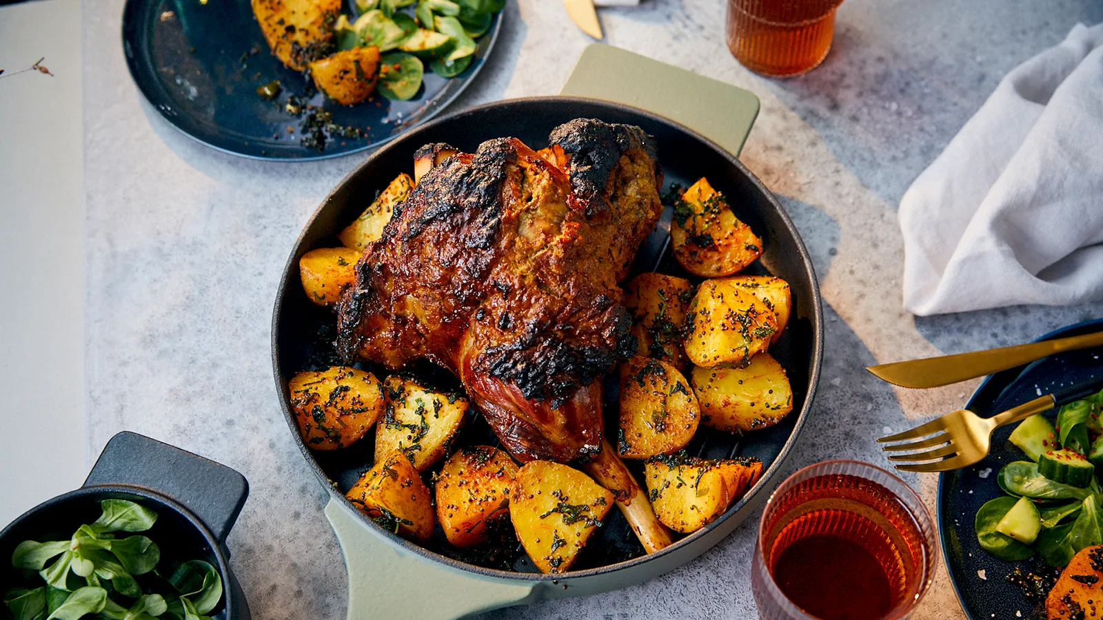 An overhead view of a roasted lamb leg with crispy skin and golden potatoes, garnished with herbs, served in a light green pan, with side salads and drinks on a grey table.