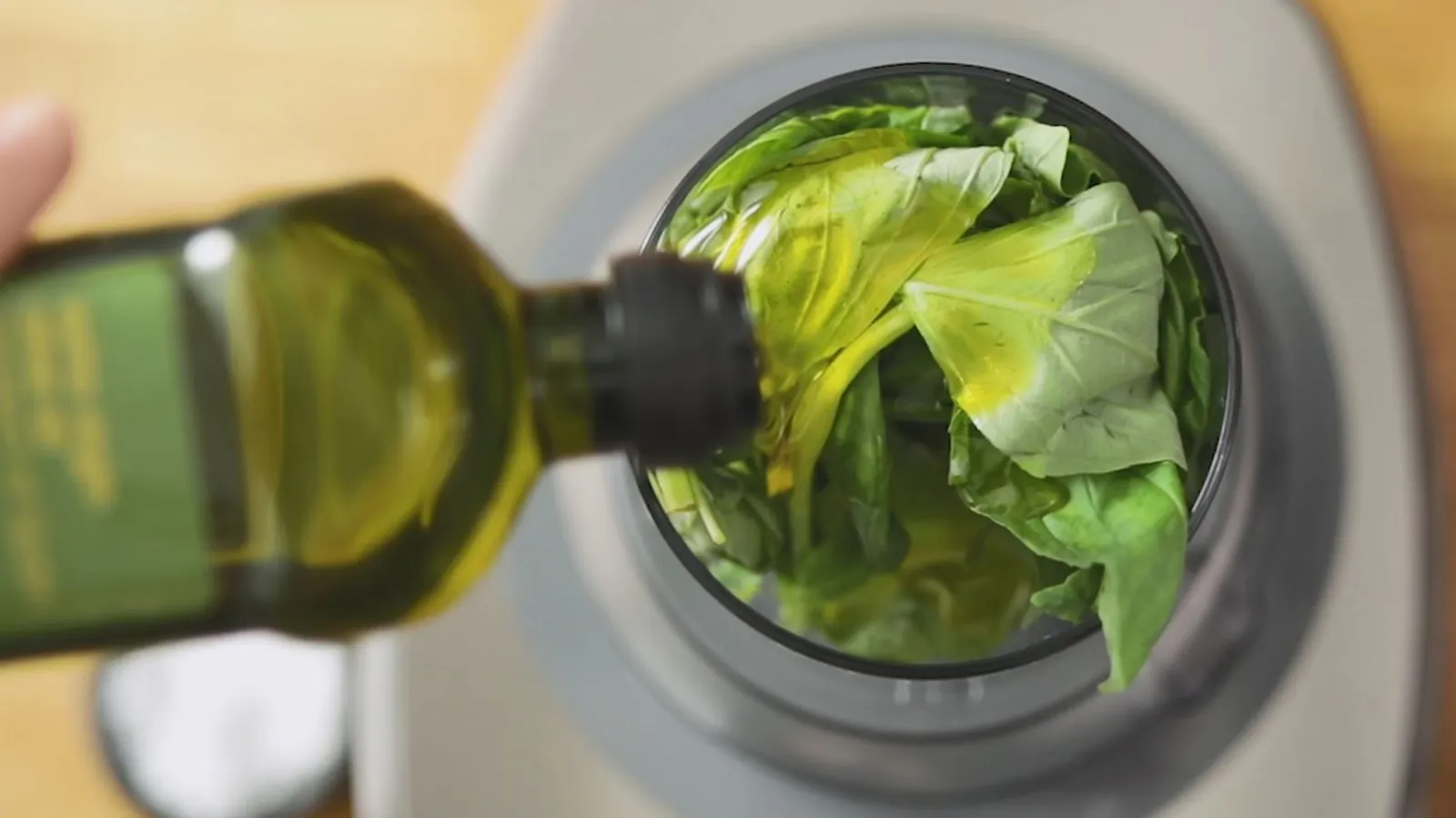 A hand pours olive oil from a green bottle into a food processor filled with fresh green basil leaves.