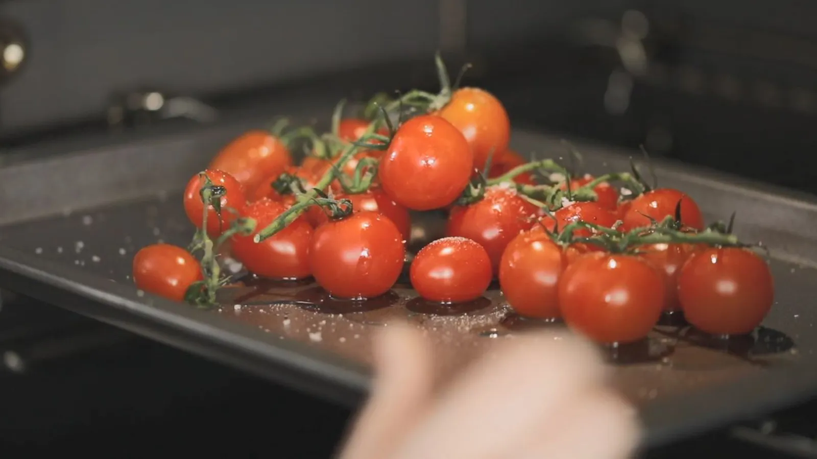 A hand slides a baking tray of fresh cherry tomatoes on the vine, seasoned with salt, into a hot oven.