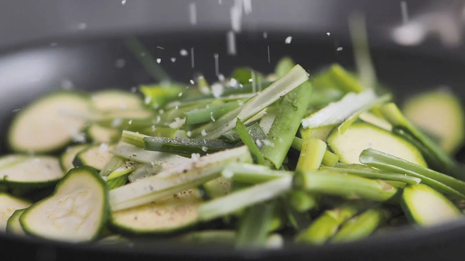 Salt is sprinkled over sliced green zucchini and green onions cooking in a black frying pan.