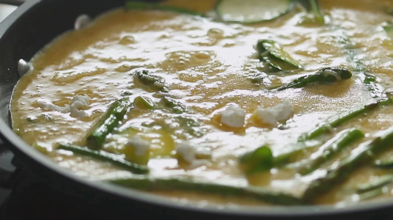 A close-up of a frittata cooking in a black frying pan, showing eggs, sliced zucchini, green beans, and small pieces of white cheese bubbling.