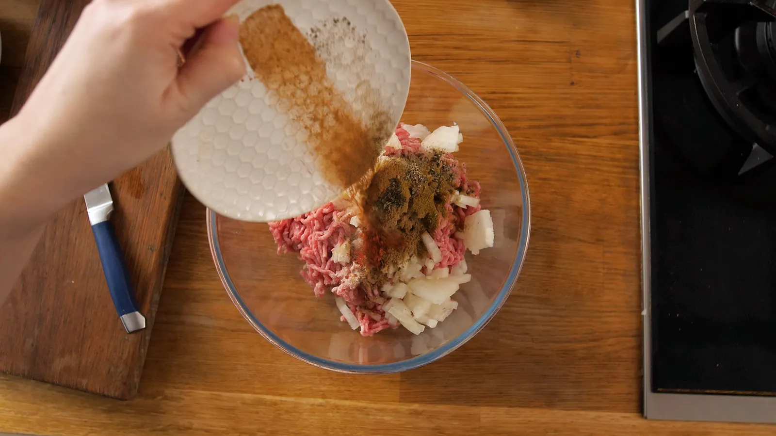 A hand pours a mixture of spices from a white plate into a clear glass bowl containing ground meat and diced onions.