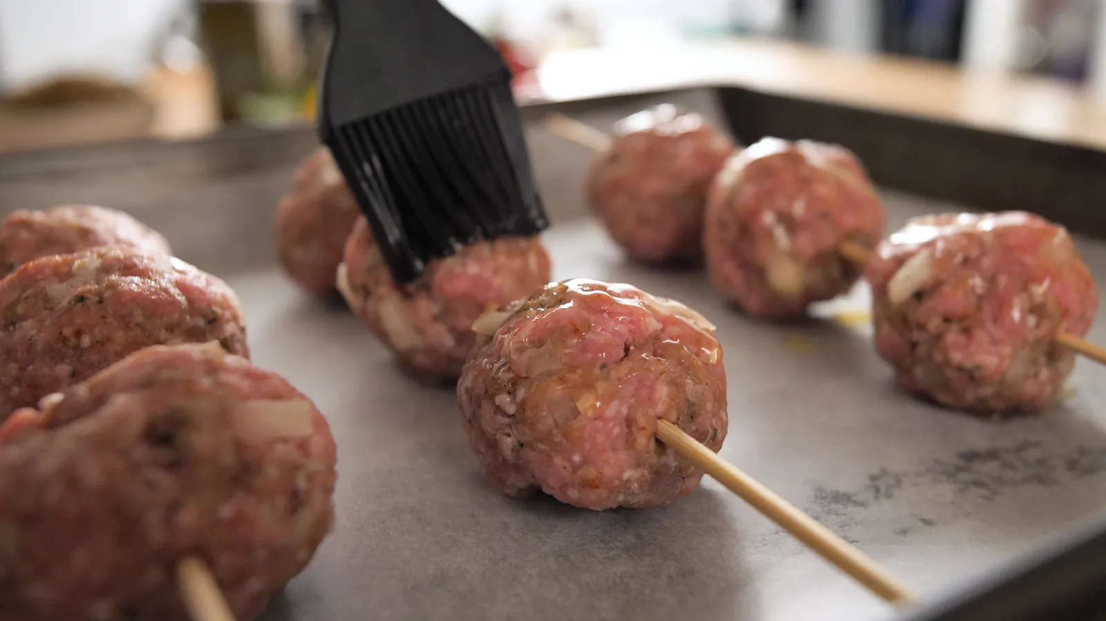 A black pastry brush glazes raw meatballs on wooden skewers, arranged on a baking tray lined with parchment paper.